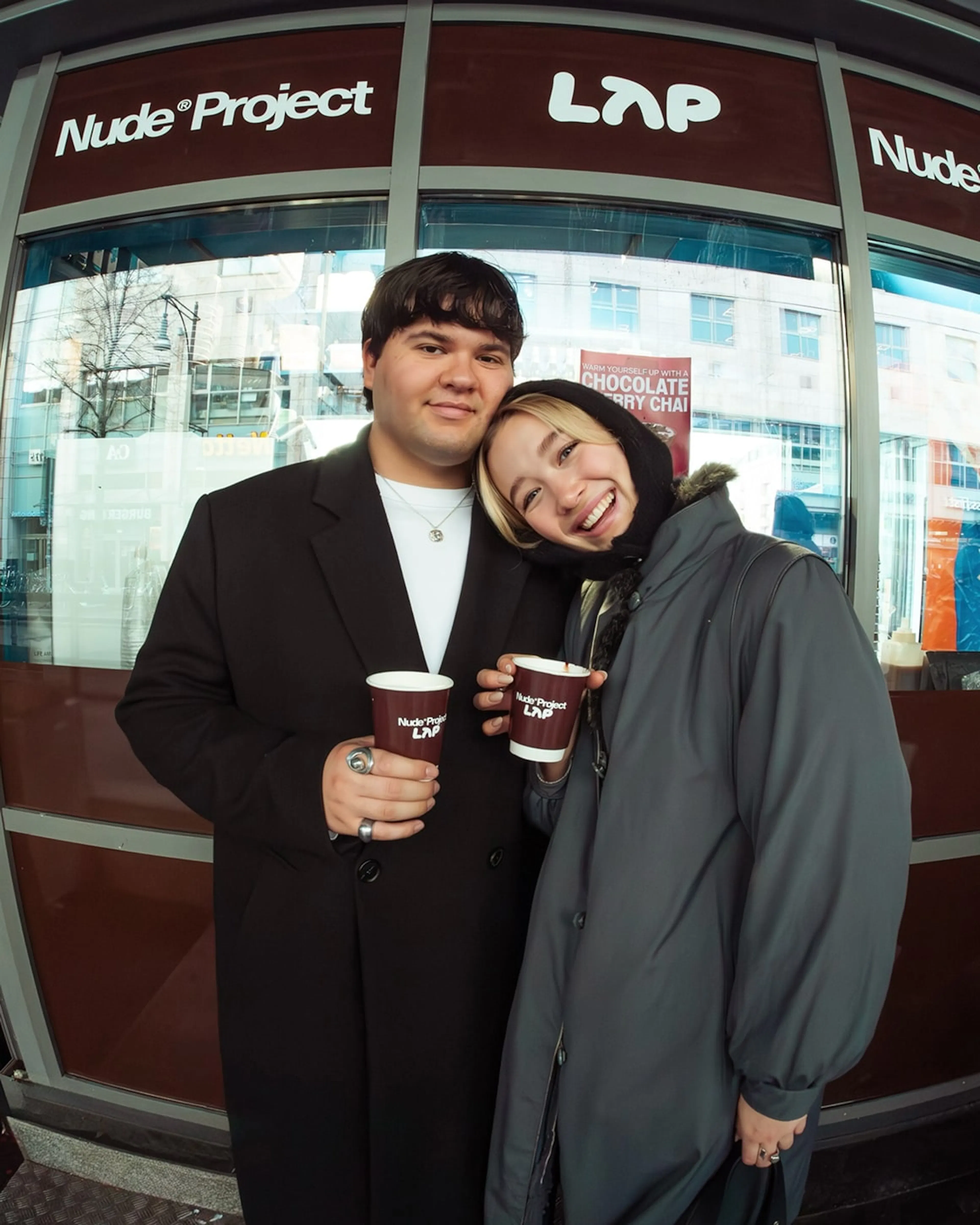 Nude Project - Communications : Photo showingTwo people holding branded coffee cups outside a Nude Project x LAP pop-up, celebrating the collaboration in a casual street setting.