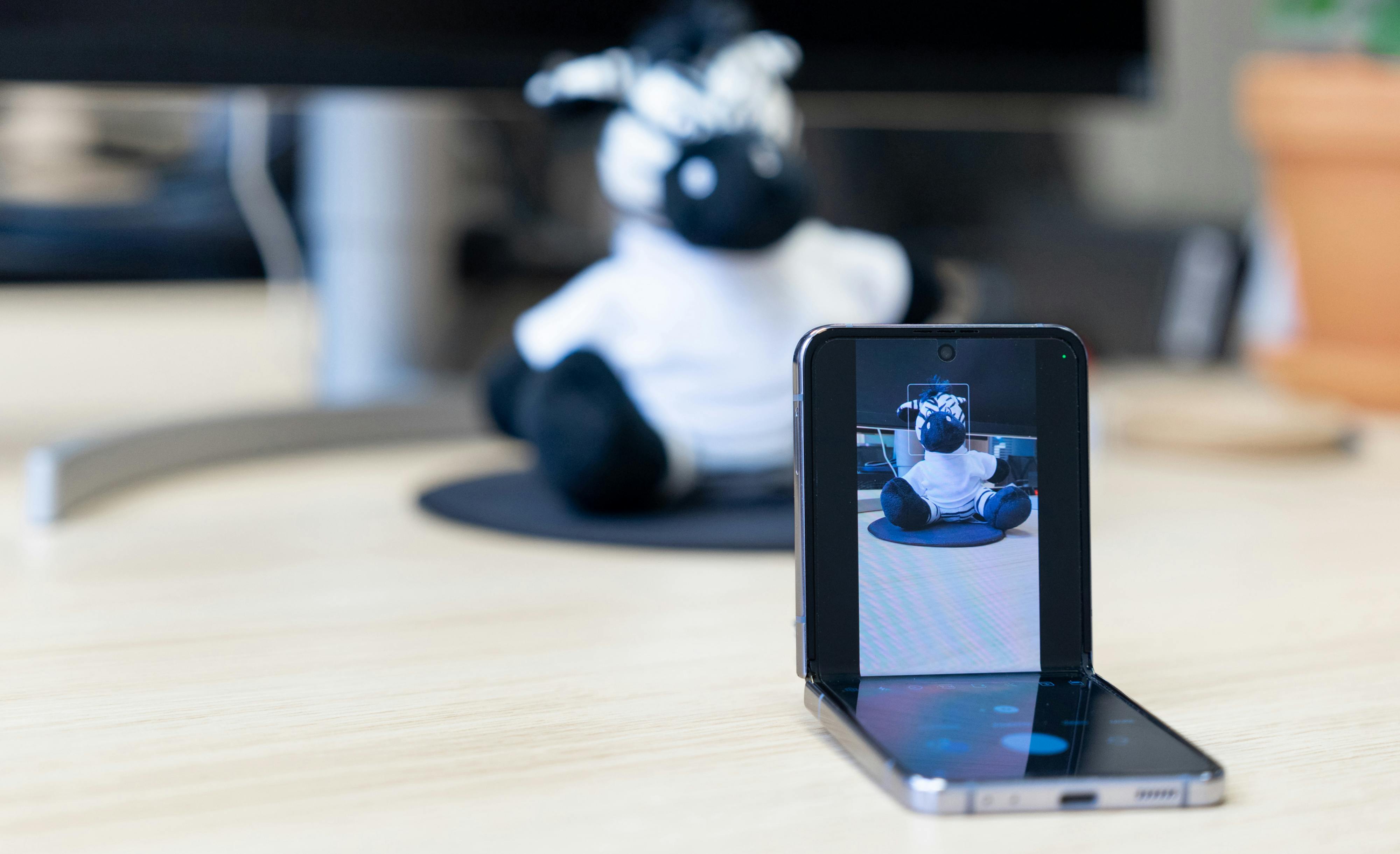 A foldable device half-folded on a desk, displaying a camera taking a picture of a plush zebra toy sitting in the background, illustrating the Flex Mode feature.