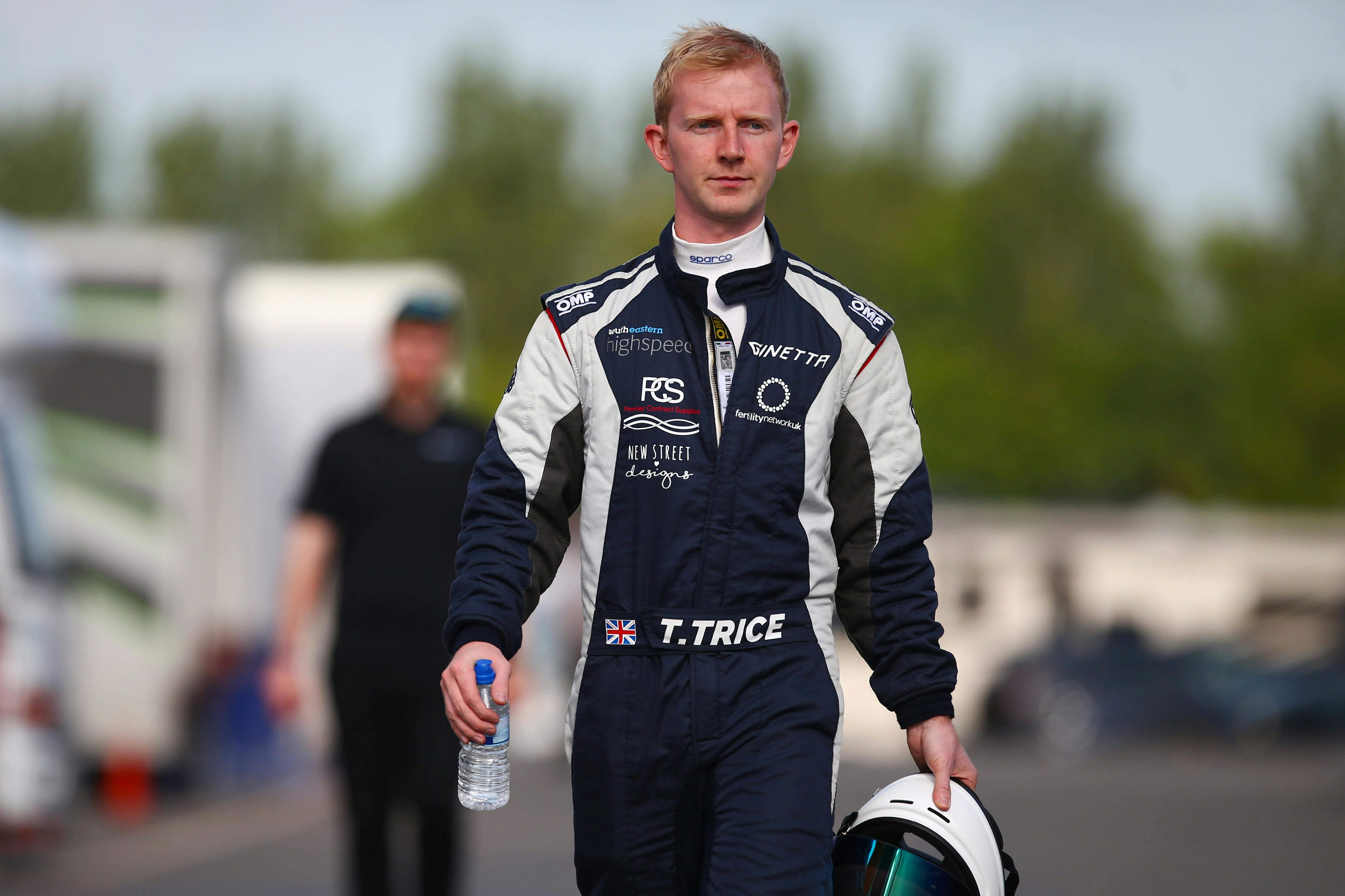 Toby Trice in his racing gear, holding the helmet in his hand, bottle of water in another, walking towards the camera.