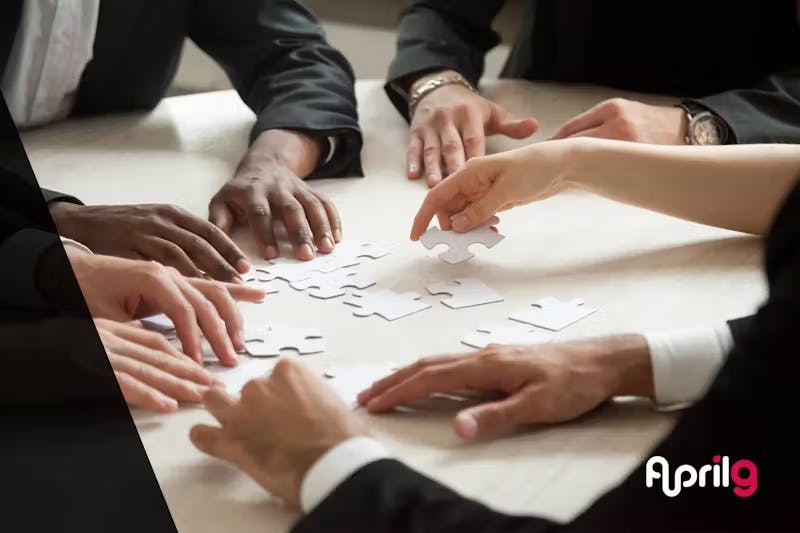 Close-up of multiple hands on a table working together to assemble puzzle pieces. The hands appear to belong to people in business attire. The April9 logo is visible in the bottom right corner.