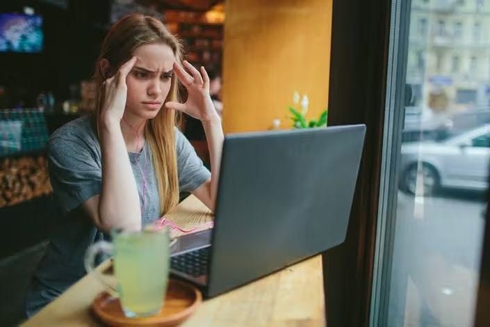 A young woman sits at a table near a window in a café, looking frustrated while staring at her laptop screen.