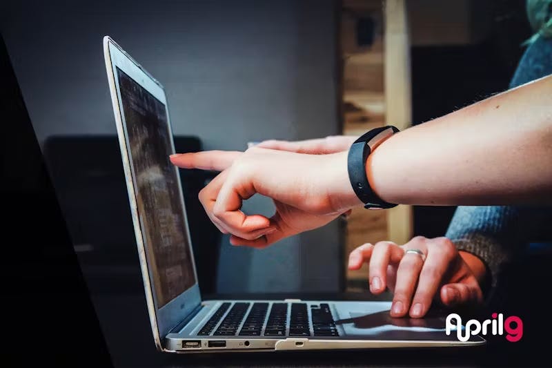 Close-up of hands using a laptop. One hand points at the screen while wearing a fitness tracker, the other rests on the touchpad. The April9 logo is visible in the bottom right corner.