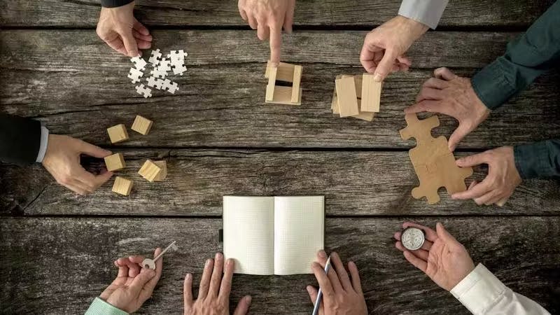 Close-up of multiple hands on a table working together to assemble puzzle pieces. 