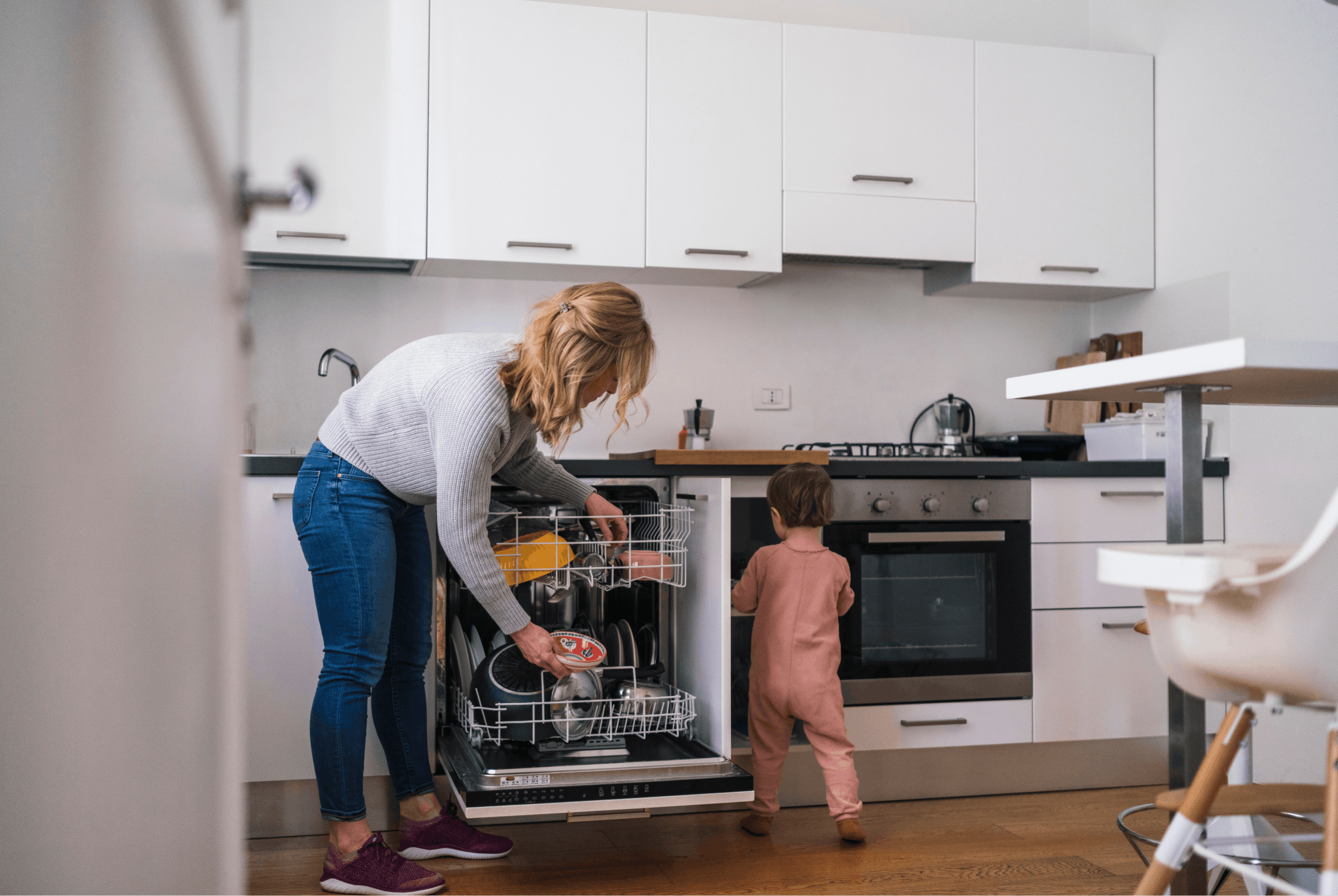 A parent is in their home's kitchen, which has white cabinets and walls. They are bent over the dishwasher, loading it with dirty dishes. Their toddler is standing next to the dishwasher.