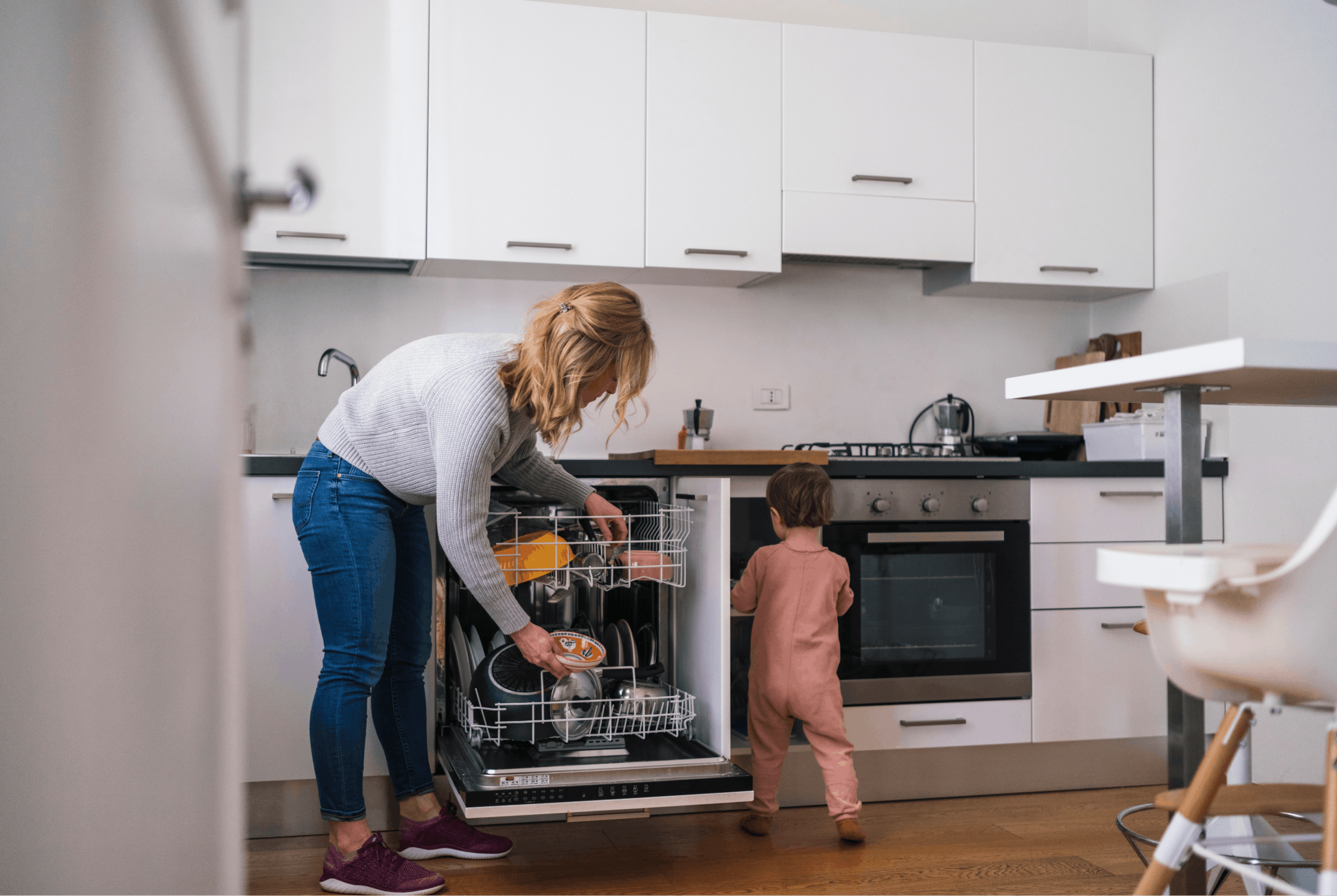 A parent is in their home's kitchen, which has white cabinets and walls. They are bent over the dishwasher, loading it with dirty dishes. Their toddler is standing next to the dishwasher. 