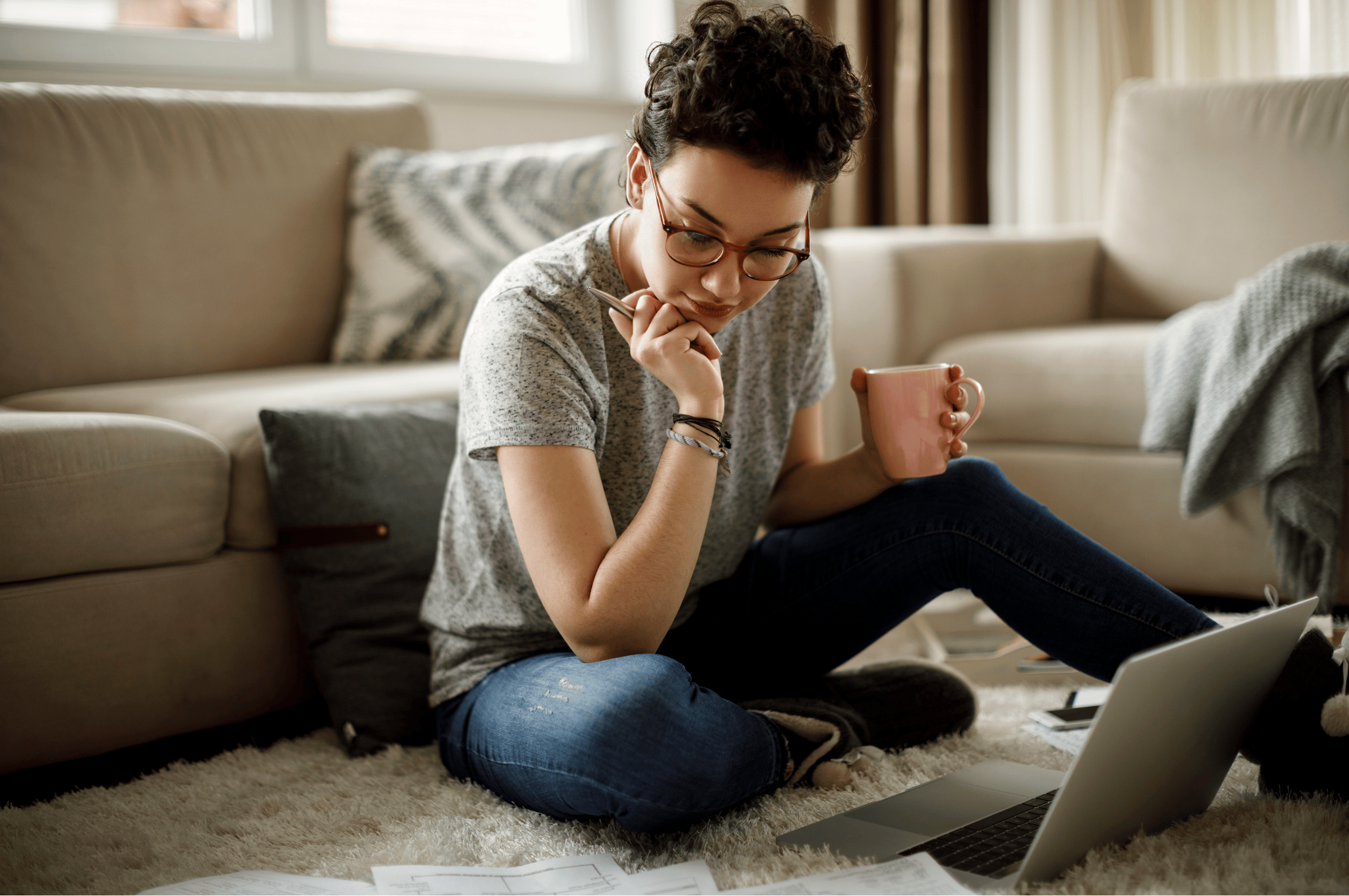 Photo of a person who is wearing glasses, a gray t-shirt, and jeans. Their hair is tied up in a bun, and they are holding a mug in one hand and a pen in the other hand. They are sitting on the floor, which is covered in a cozy, white rug. They are looking down at some papers, presumably bills, while their laptop sits open near them.