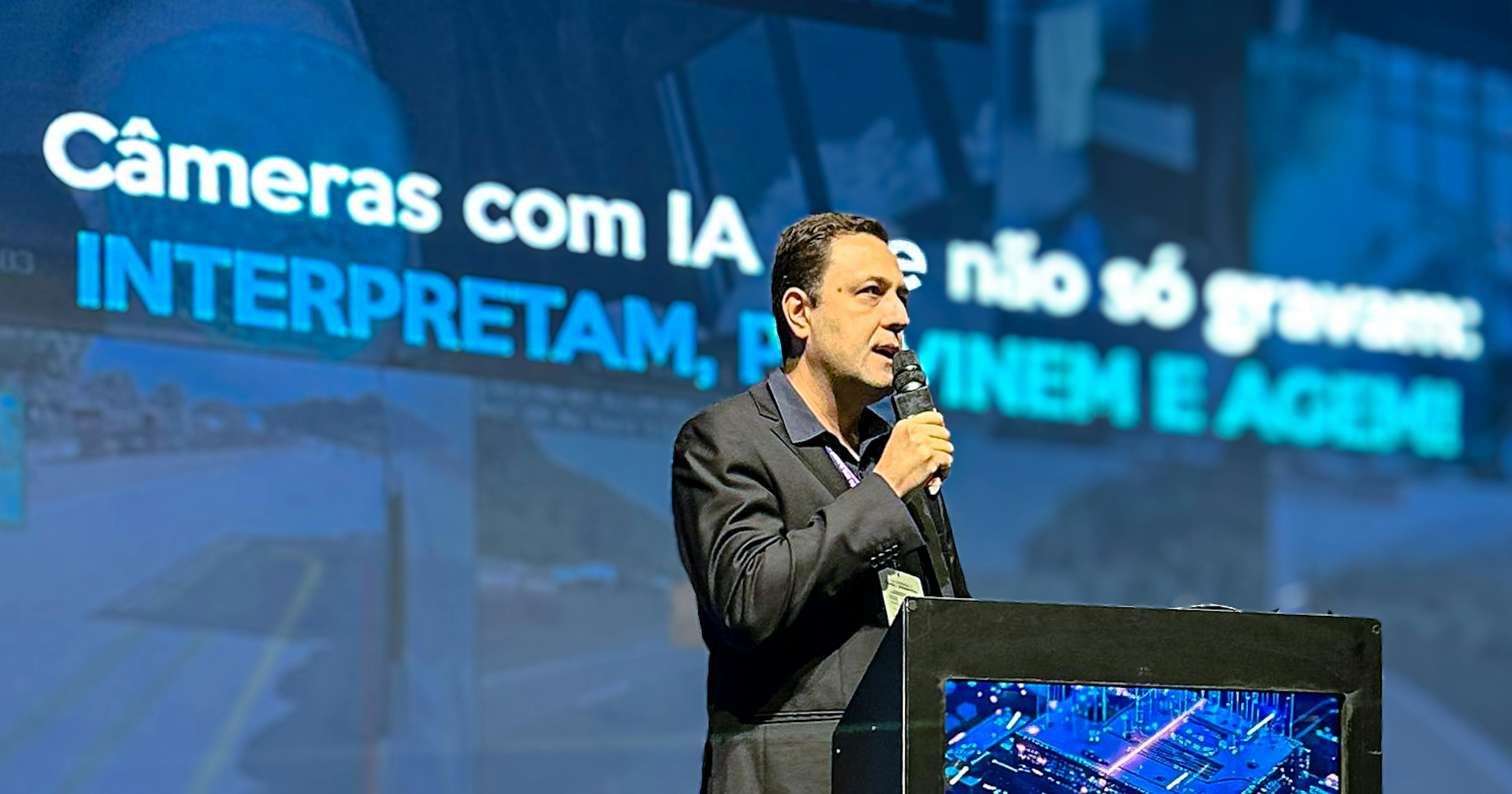 Homem de terno escuro fala ao microfone em um palco durante evento, com um telão ao fundo exibindo a frase “Câmeras com IA não só gravam: INTERPRETAM, PREVINEM E AGEM!”. A iluminação destaca o apresentador, enquanto o restante do ambiente permanece em tons azulados e tecnológicos, transmitindo uma atmosfera de inovação.