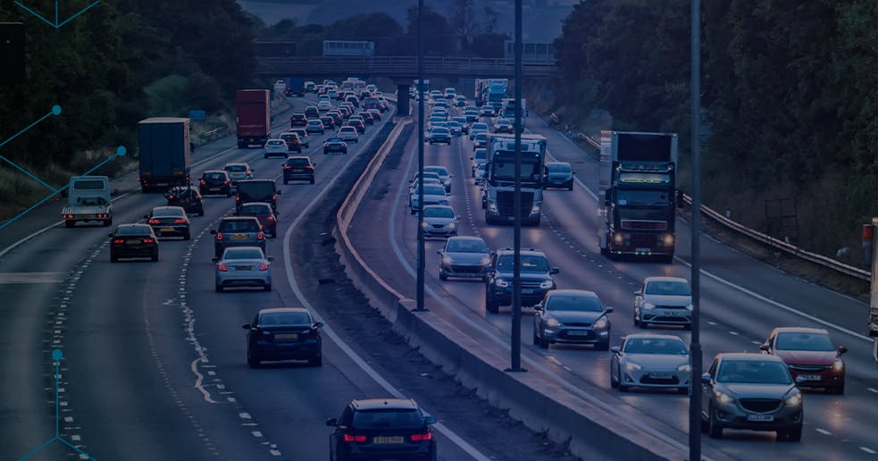 Rodovia movimentada com fluxo intenso de carros e caminhões em várias faixas, cercada por árvores, com sobreposição de gráficos em forma de hexágonos azuis que remetem à tecnologia e conectividade.