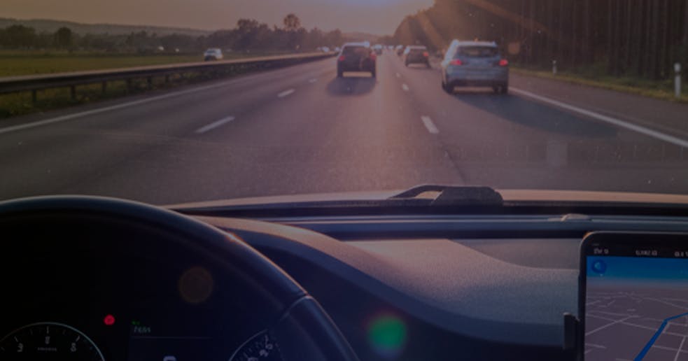 Visão do interior de um carro em movimento durante o pôr do sol, mostrando o volante, o painel e um celular com aplicativo de navegação ligado, enquanto o veículo percorre uma rodovia com outros carros à frente.