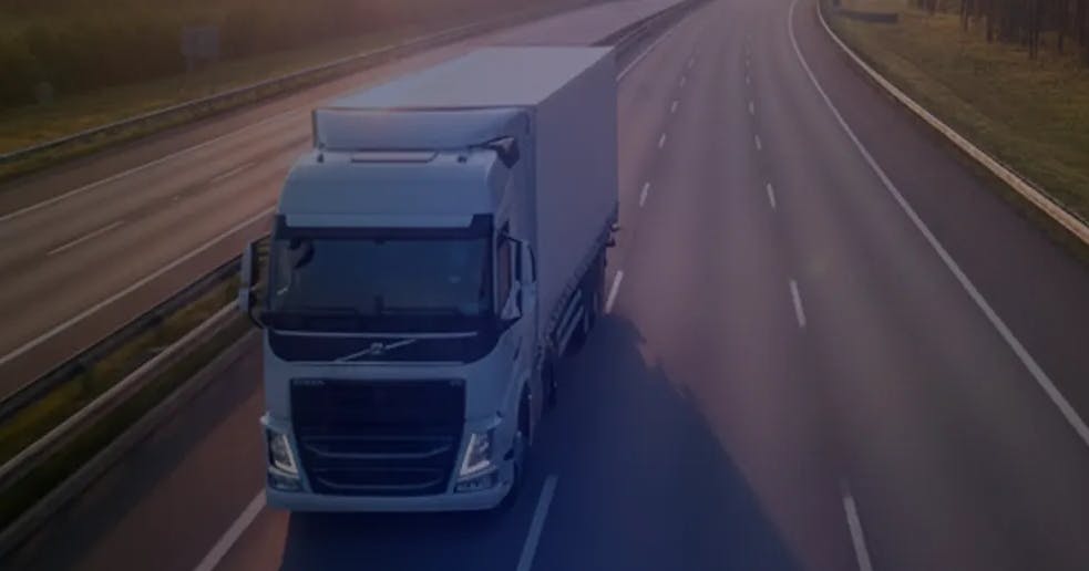 Caminhão trafegando em uma rodovia durante o pôr do sol, com poucas faixas de veículos e vegetação ao redor, representando transporte rodoviário e logística.