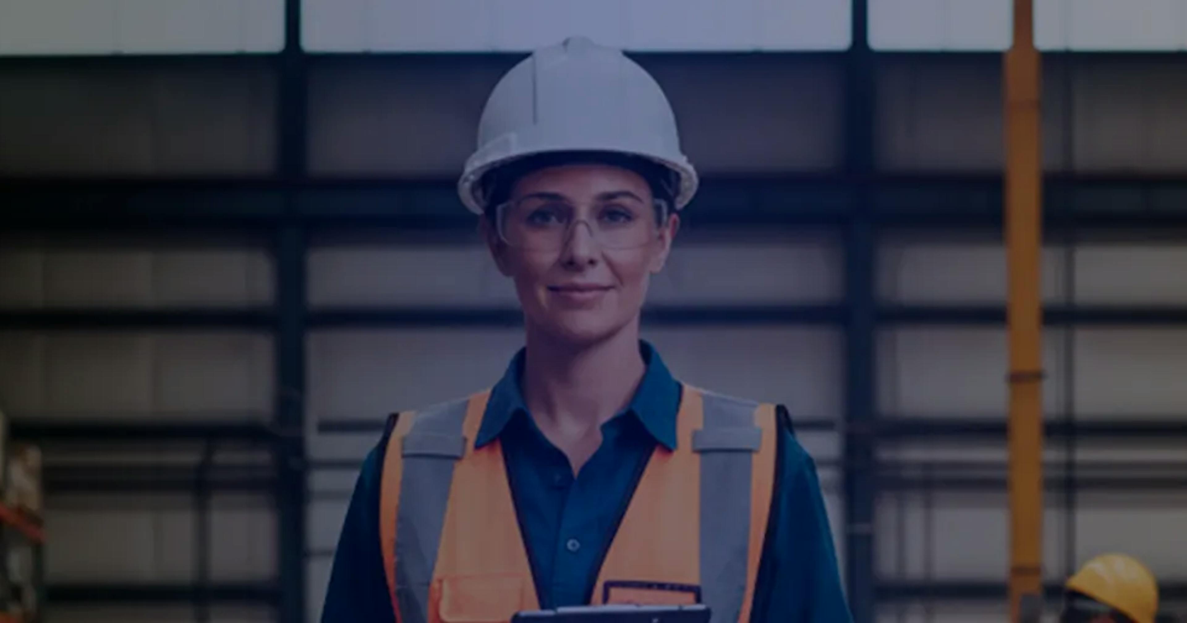 Mulher usando capacete de segurança, óculos de proteção e colete refletivo dentro de um armazém industrial. Ela está de pé, segurando uma prancheta, com prateleiras de estoque desfocadas ao fundo.