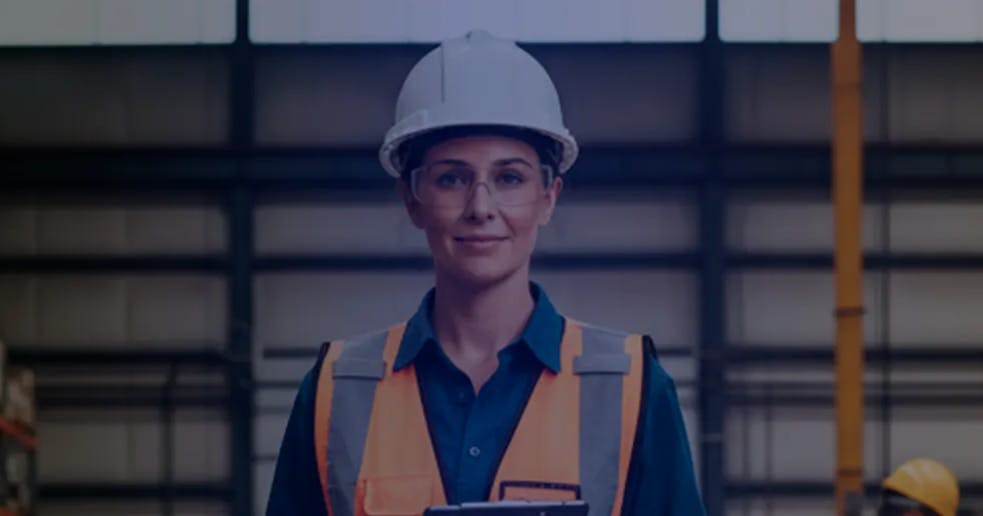 Mulher usando capacete de segurança, óculos de proteção e colete refletivo dentro de um armazém industrial. Ela está de pé, segurando uma prancheta, com prateleiras de estoque desfocadas ao fundo.