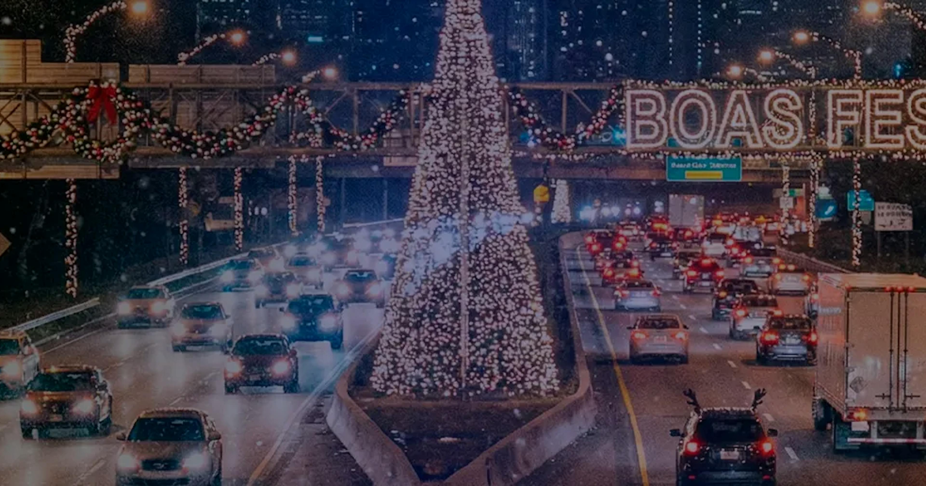 Rodovia movimentada à noite decorada com luzes e enfeites de Natal, incluindo uma grande árvore iluminada no canteiro central e uma faixa luminosa com a mensagem 'Boas Festas' sobre as pistas.
