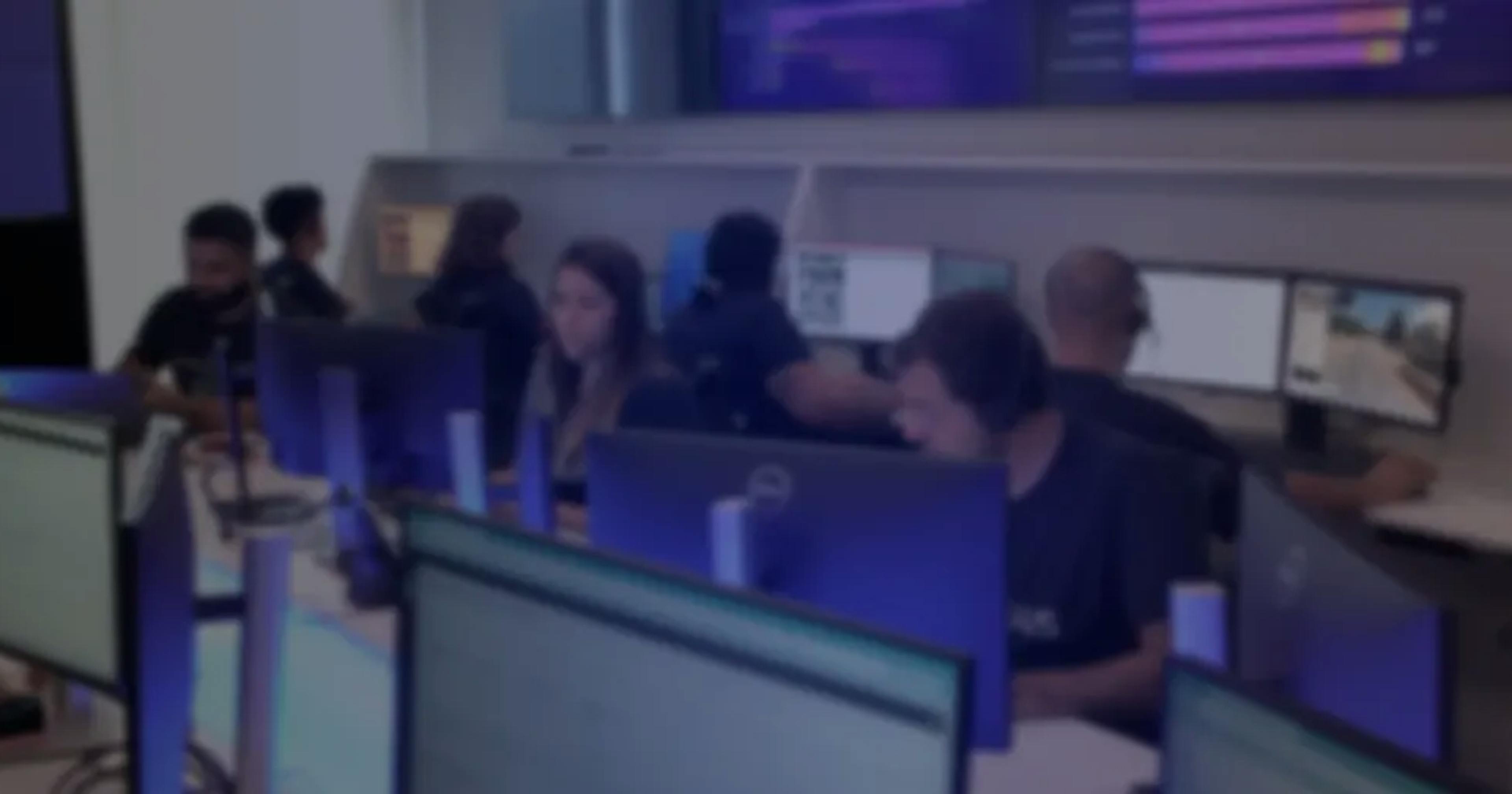 Equipe trabalhando em uma central de operações, sentada em estações com múltiplos monitores exibindo gráficos e imagens de vídeo.