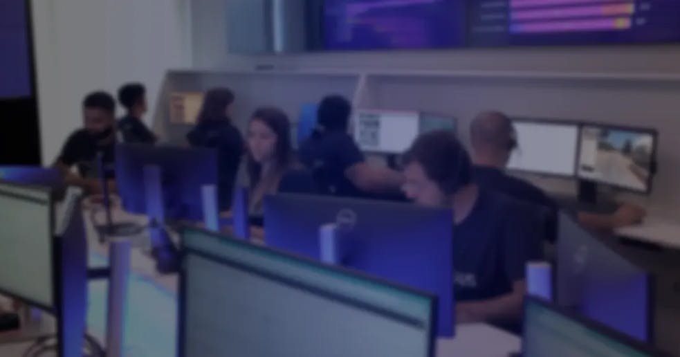 Equipe trabalhando em uma central de operações, sentada em estações com múltiplos monitores exibindo gráficos e imagens de vídeo.