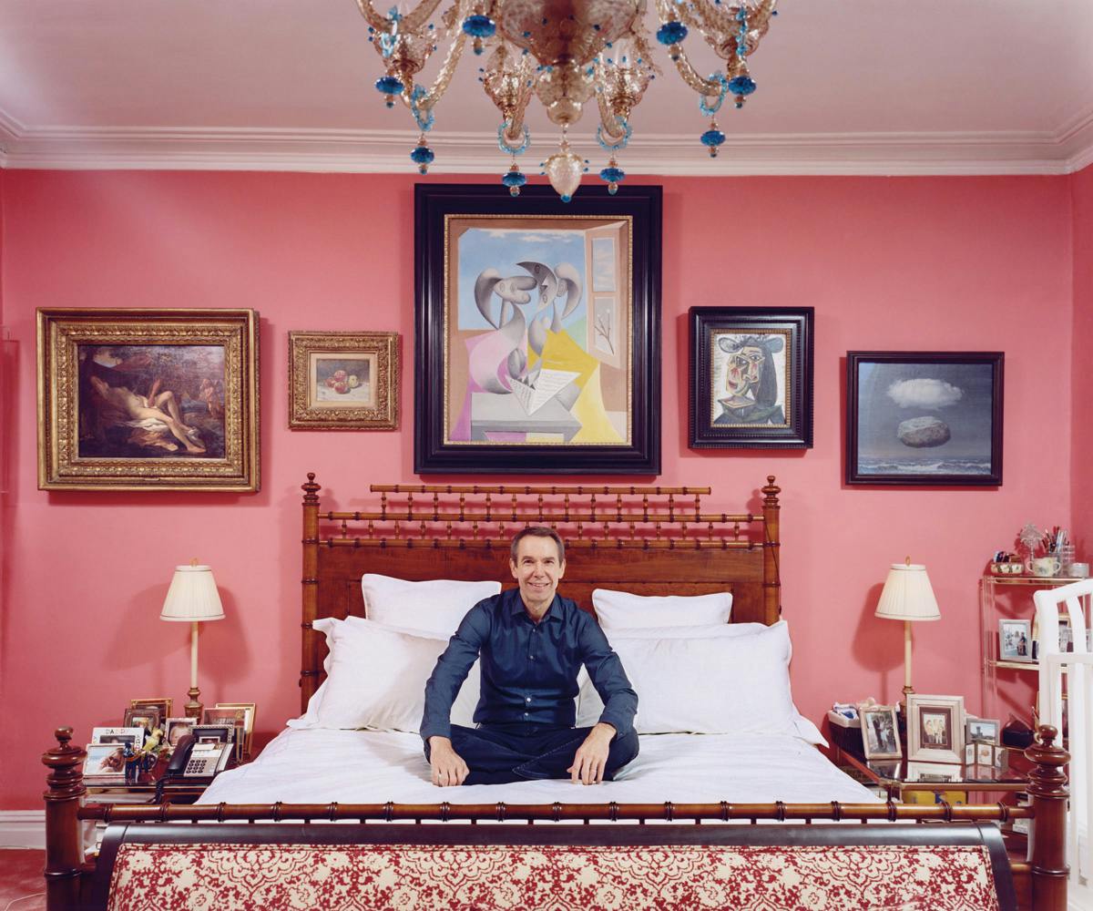 Jeff Koons sitting on his bed in front of his art collection.
