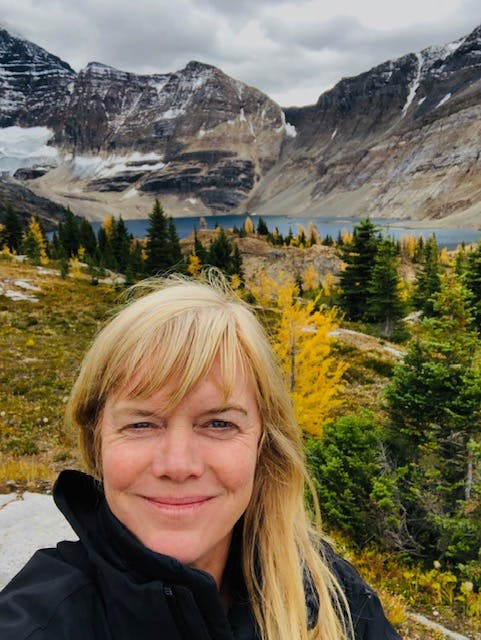The image features a person, Adrienne Lawlor, smiling with trees and mountains in the background, set against a sky with clouds. The person appears to be posing outdoors in a natural setting during fall, with snow visible on the mountains.