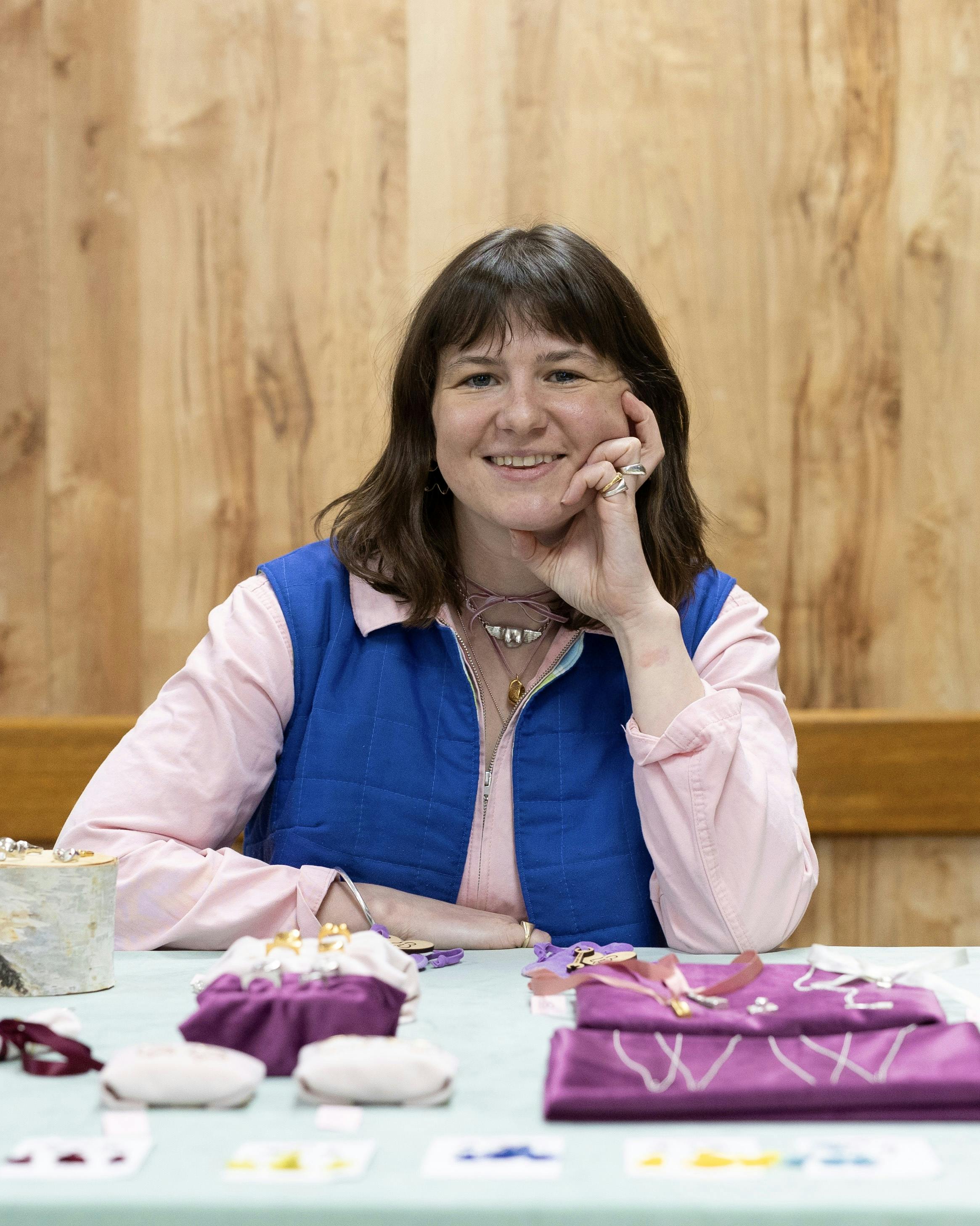The image features a woman, Gini Dickenson, sitting at a table indoors. She is wearing purple clothing and is smiling. The background includes a wooden wall. 