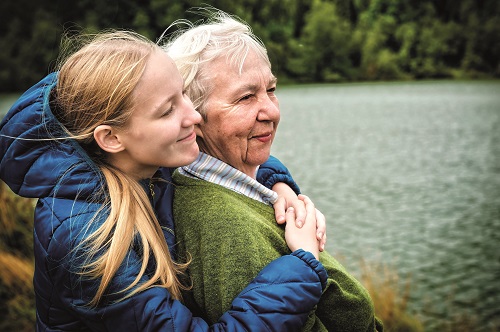 A younger woman standing behind an older woman and hugging her. Both are smiling and there is a lake behind them.