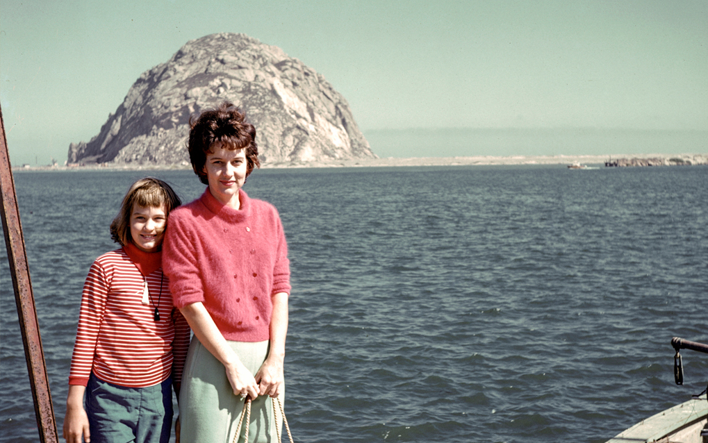 A girl and woman, both wearing pink tops, standing on the deck of a boat and looking straight at the camera. There is a big rock in the background.