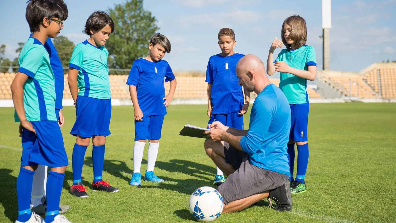 Youth coach kneeling with a clipboard in front of his team on the football pitch