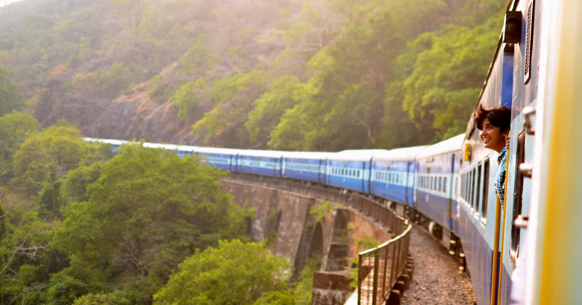 Blue train on the edge of a mountain with green trees.