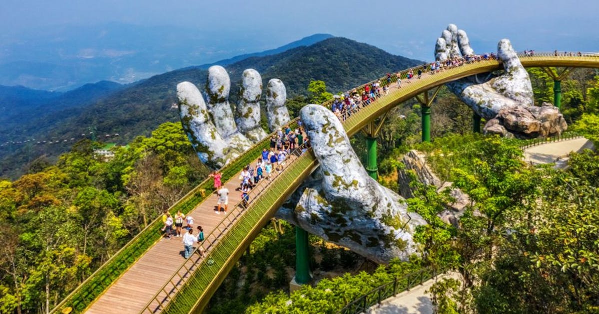 Aerial photo of a bridge held up by two giant stone hands. The bridge is surrounded by greenery.