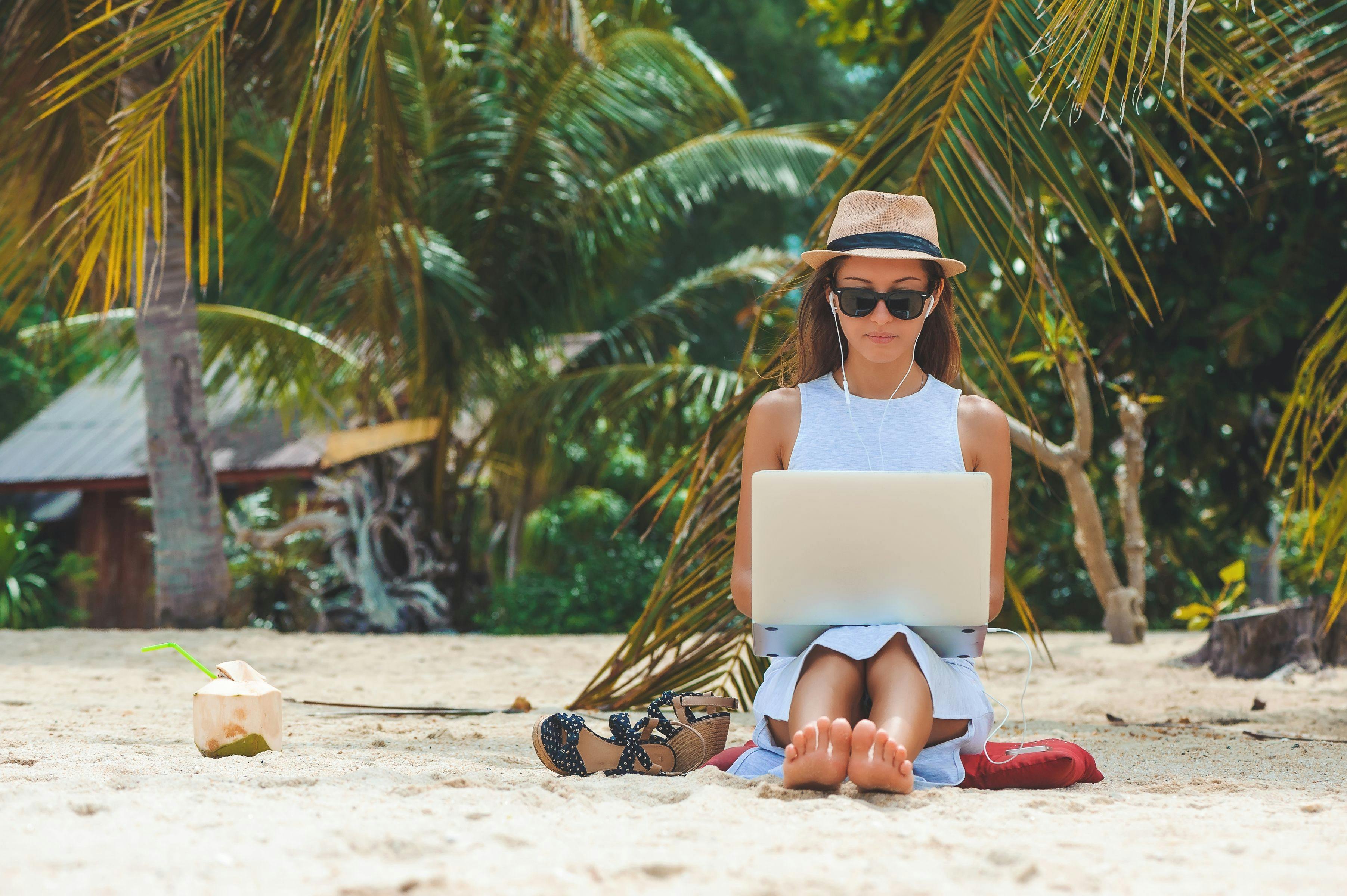 A person sitting on a beach while busy on their laptop with palm trees and a wooden building in the background.