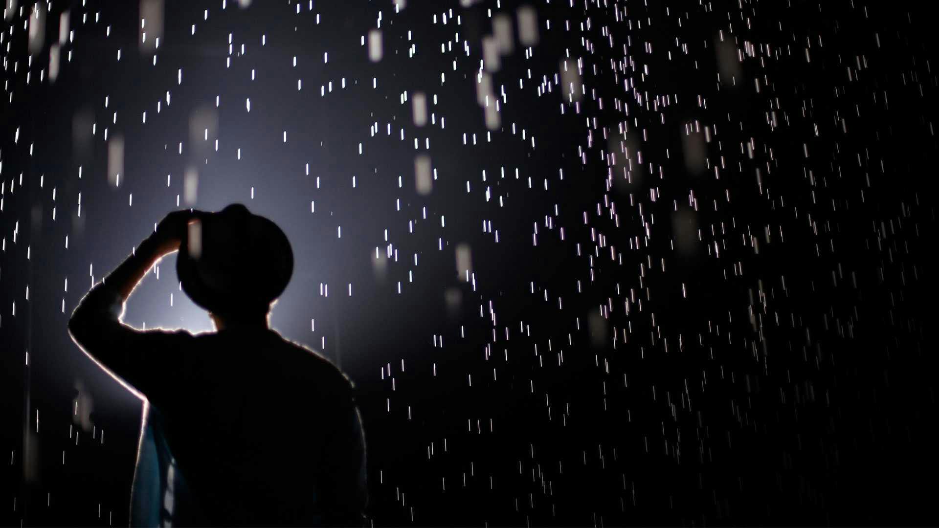 A dark picture with visible rain drops while a person is holding their hat in place.