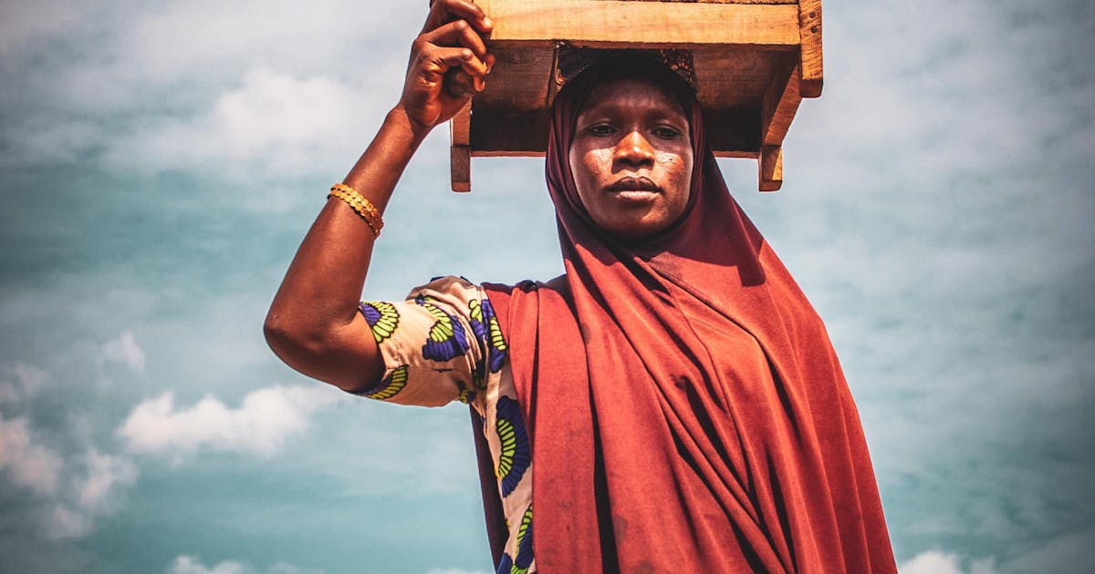 A photo of a Nigerian woman carrying a box on her head on a slightly cloudy day in Nigeria.