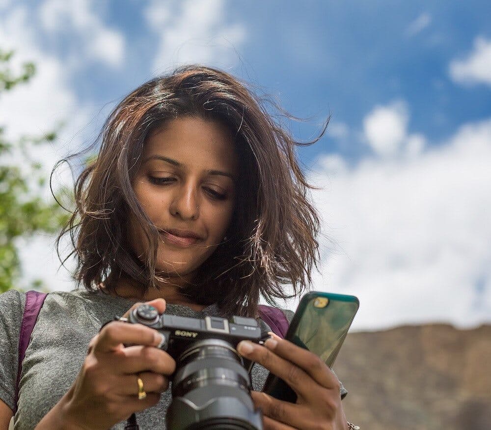 Photo of a woman looking down at her camera while holding her phone during daytime.
