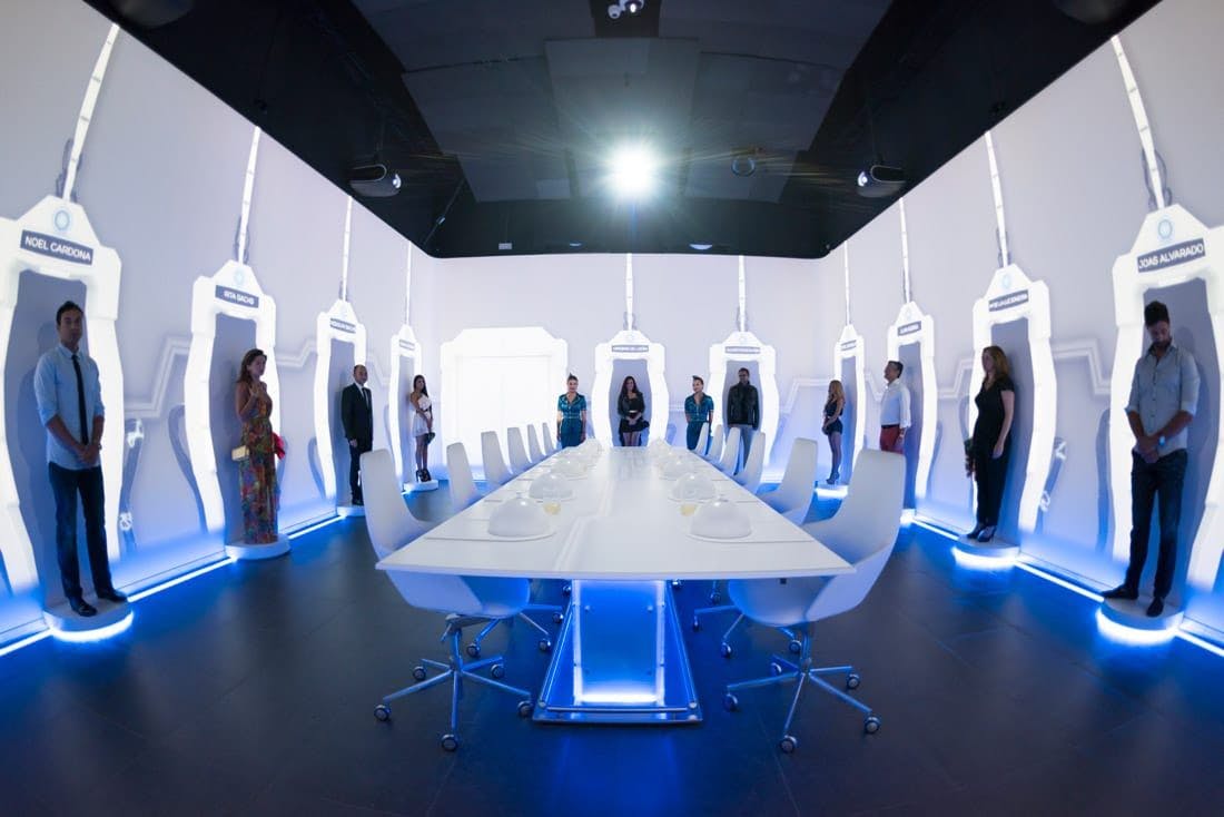 A room with a white table and chairs in it, while people are standing against the walls.