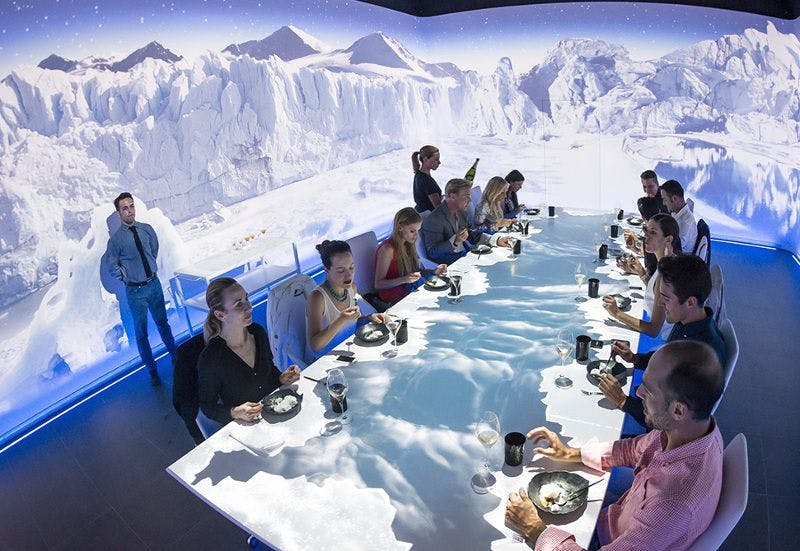 Multiple people sitting around a table containing food and drinks with a background of mountains covered in ice.