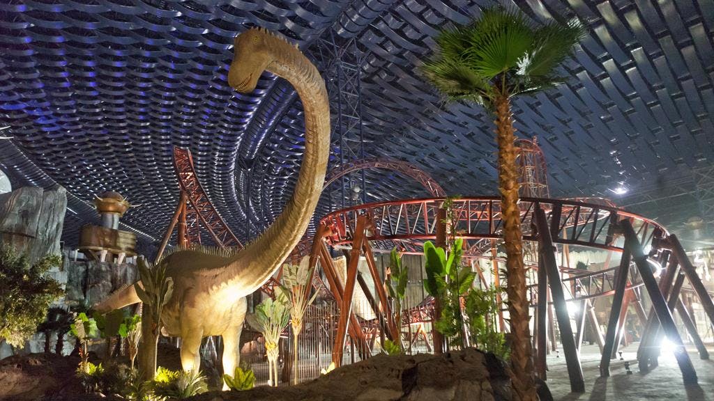 A palm tree and a replica of a dinosaur next to a roller coaster structure.