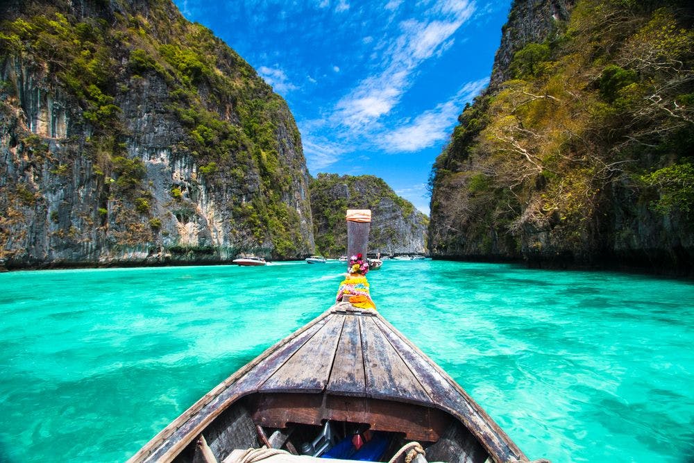 First person view of a wooden boat driving on crystal clear blue water between two big island rocks.