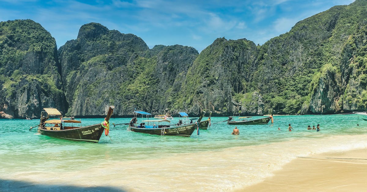 Far away photo of long boats floating on clear blue water surrounded by grassy mountains.