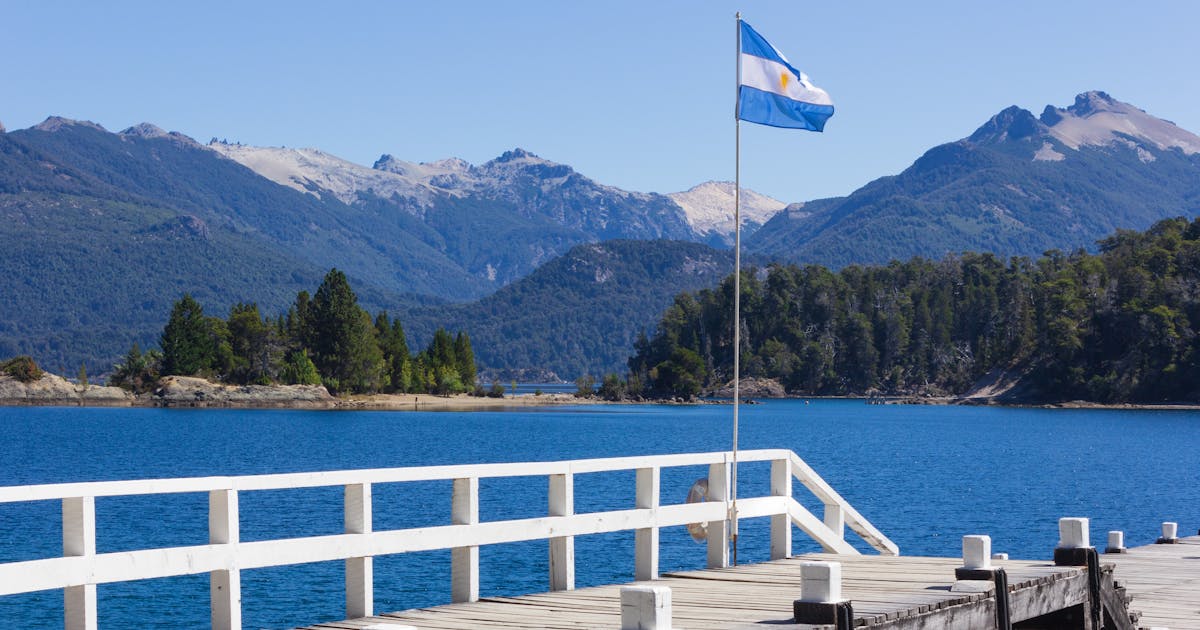 Photo of a blue and white flag waving in the wind on a pier, surrounded by blue water and mountains.