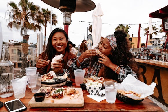 Two women eating at a table outdoors and smiling.