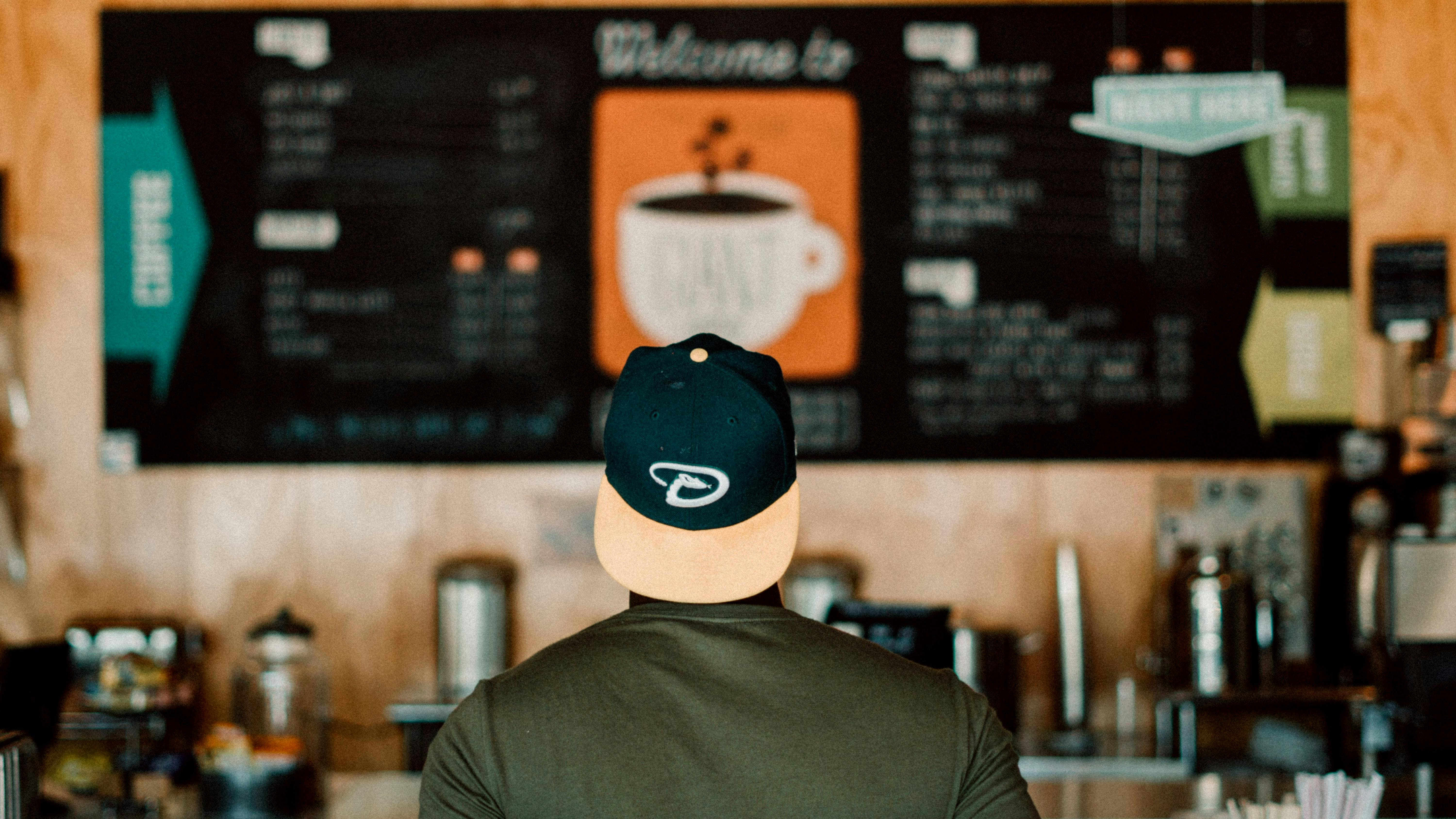 The back of a man with a hat looking at the menu in a coffee shop. 