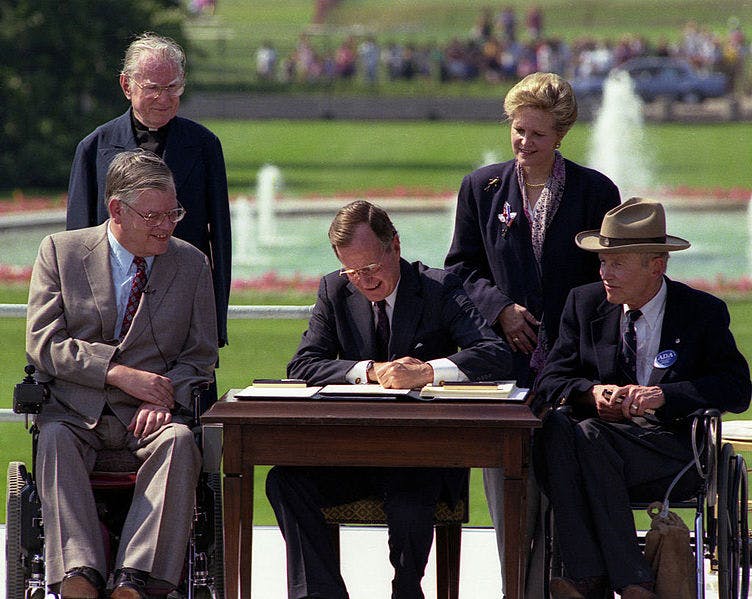 On July 26, 1990, President Bush signs the Americans with Disabilities Act on the South Lawn of the White House. Sharing the stage with the President as he signs the Act are (standing left to right): Rev. Harold Wilkie of Claremont, California; Sandra Parrino, National Council on Disability; (seated left to right): Evan Kemp, Chairman, Equal Opportunity Commission; and Justin Dart, Presidential Commission on Employment of People with Disabilities. Mrs. Bush and Vice President Quayle participate in the Ceremony.
