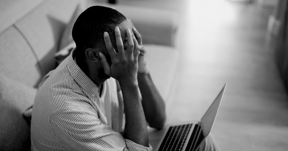 Image a black man frustratingly holding his head in his hands while looking at a laptop