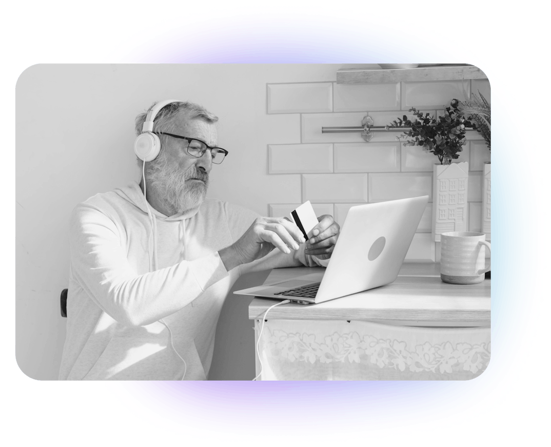 A senior man with glasses and headphones on sitting at a laptop and holding a credit card