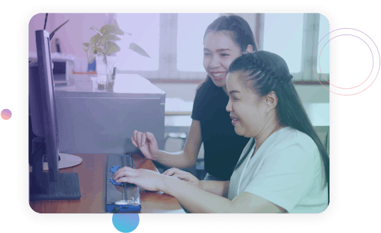 An asian woman who is blind using a braille display for her computer. Another woman is sitting next to her smiling.
