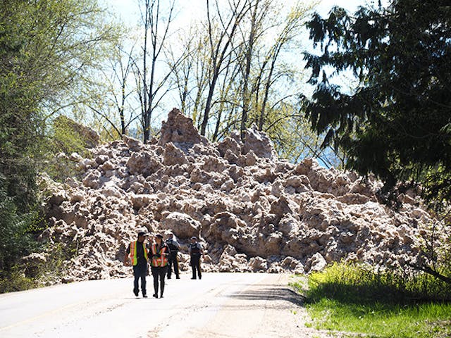 A pile of wet snow avalanche debris covers a road up to 10 metres deep. Four people are on the road looking at it.