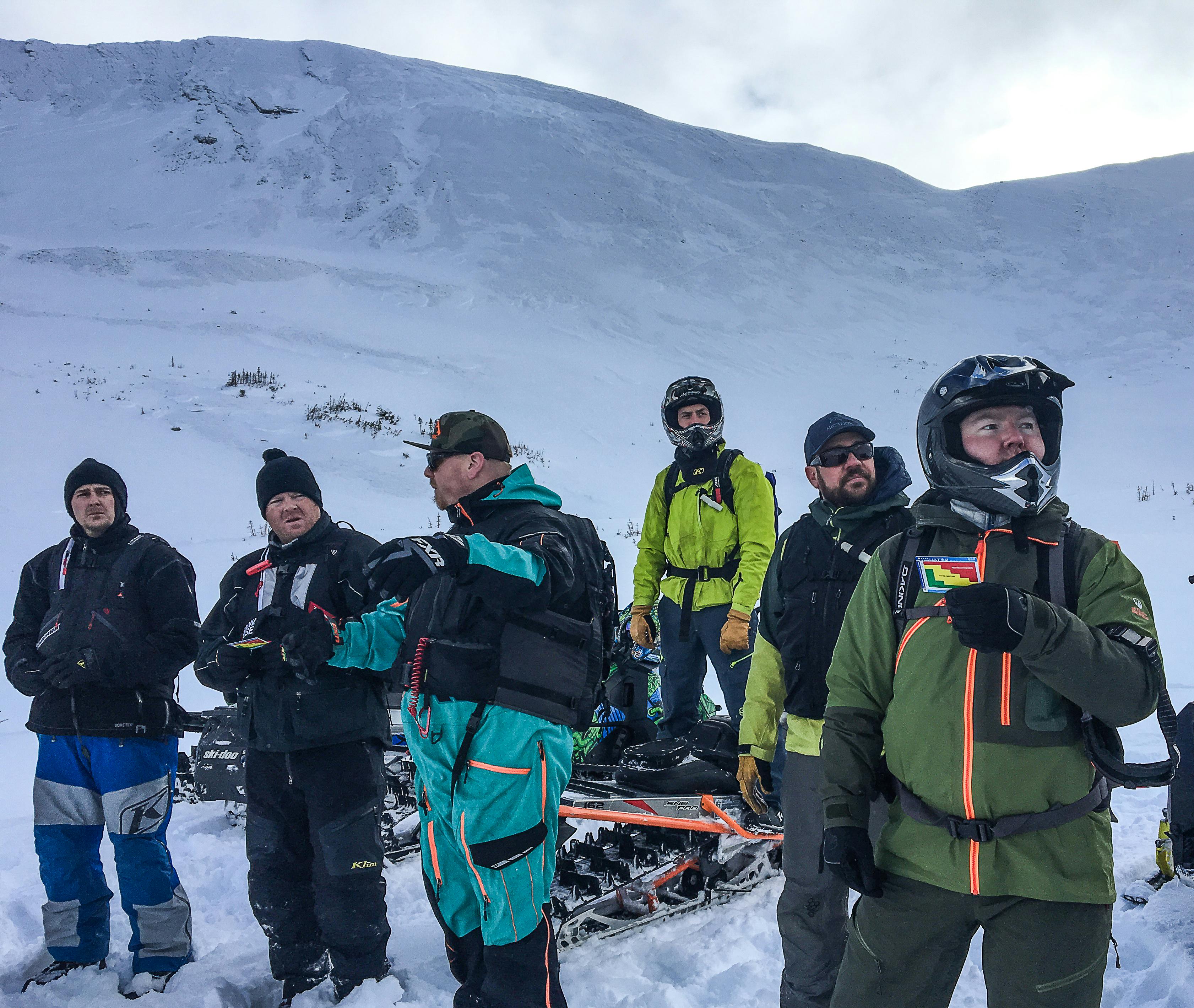 A group of sledders stand together discussing terrain. One is motioning to another with his hands as he talks and another is using an Avaluator card.