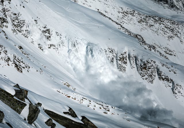 A large avalanche in motion over some cliffs. On the slope above it is a person standing and watching (maybe the trigger) and some ski tracks. 