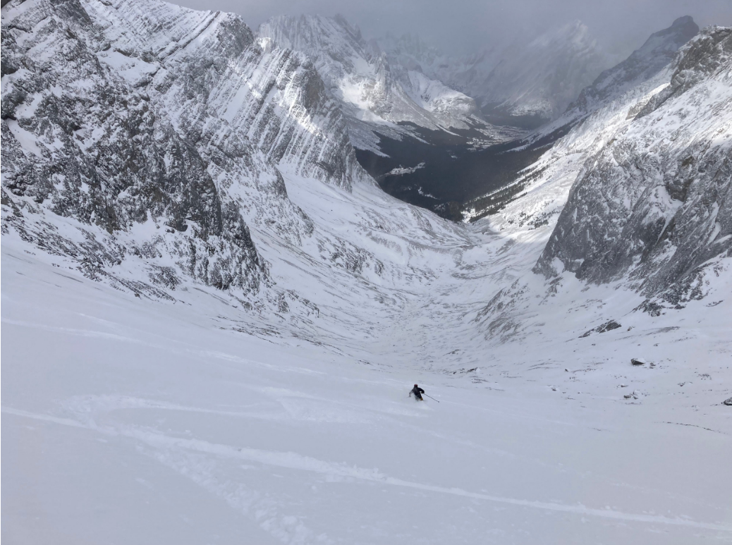 A skier rides down a slope into the valley. Fresh snow covers the slope and there are some other tracks.