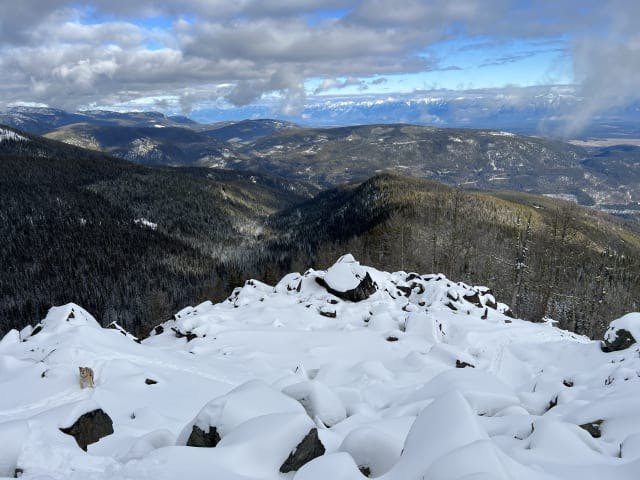 Snow barely covers the rocks in a boulder field, in the distance are other peaks with limited snow cover. In the bottom left of the image, there is a dog walking along a path in the snow. 
