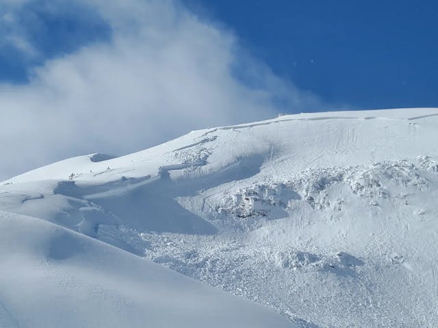 An alpine bowl shaped feature. An avalanche has propagated across the whole ridgeline. There are cliffs visible in the avalanche path and hangfire at the top. The sky is blue and it is sunny. 