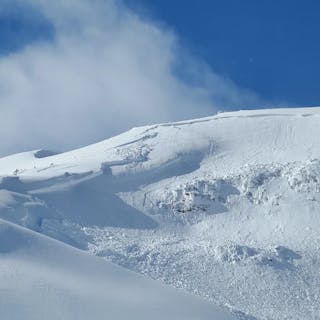 An alpine bowl shaped feature. An avalanche has propagated across the whole ridgeline. There are cliffs visible in the avalanche path and hangfire at the top. The sky is blue and it is sunny. 